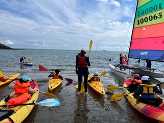 Sortie kayak en mer durant le Village Copain du Monde 2023 à Paimpol. Nombreux kayaks de dos, face à la mer, avec des enfants dedans prêts à partir. Un catamaran sur la droite.