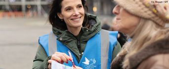 Une bénévole, souriante, du Secours populaire, propose à une passante de faire un don pour l'association.